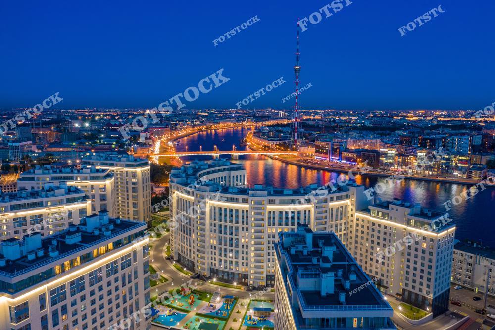 На фотографии запечатлена панорама Санкт-Петербурга с высоты птичьего полета в ночное время. В центре внимания – современные жилые комплексы на набережной реки Невы. Вдали видна знаменитая Санкт-Петербургская телебашня, которая возвышается над городом, а также ярко освещенный мост, пересекающий Неву. Город погружен в огни, отражающиеся в водах реки.
